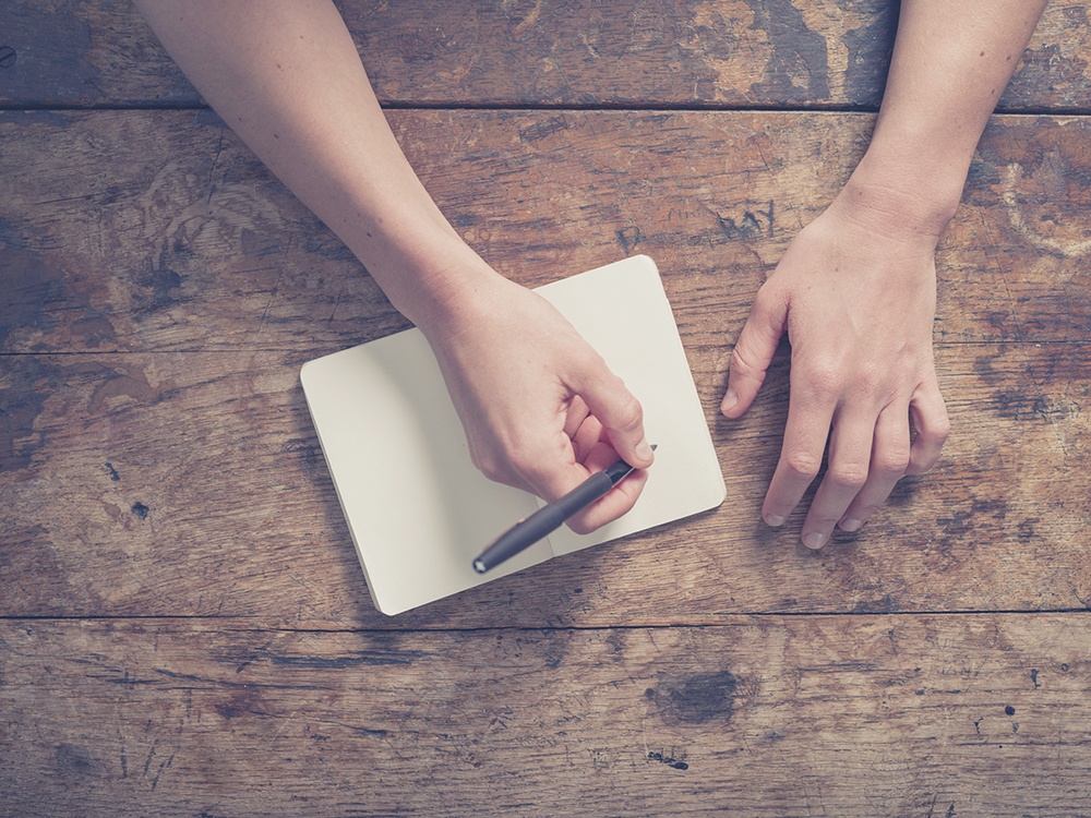 Image: iStock/Close up on the hands of a young woman as she is writing in a small notepad at a wooden table