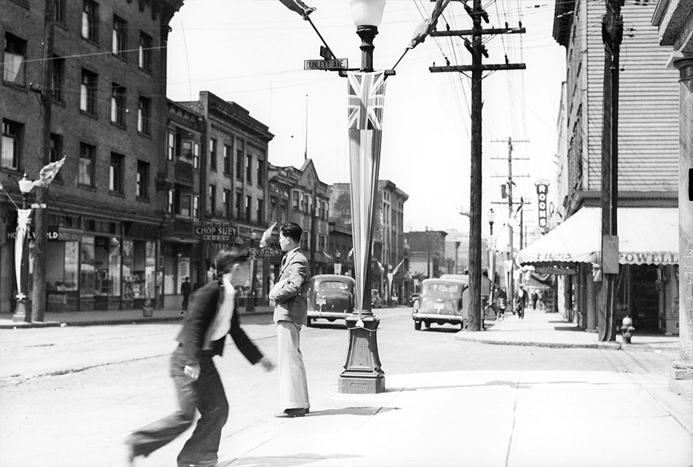 1939: Two Nikkei cross Powell Street by a lightpost emblazoned with the Union Jack. (Vancouver Archives)